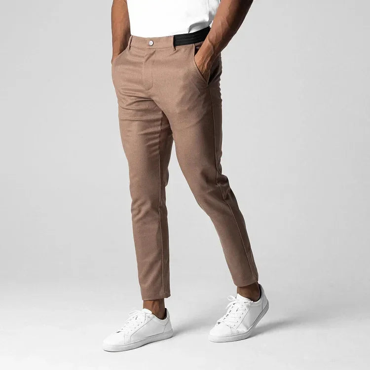 Man wearing slim-fit brown trousers with side pockets and white sneakers, standing on gray background