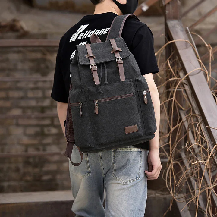 Person wearing black canvas backpack with brown leather straps standing near industrial stairs