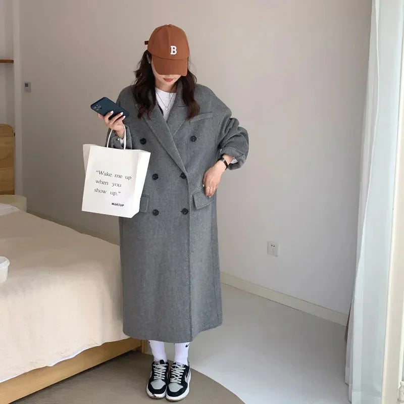 Woman wearing a long grey double-breasted coat, brown cap, and sneakers holding a shopping bag and phone indoors