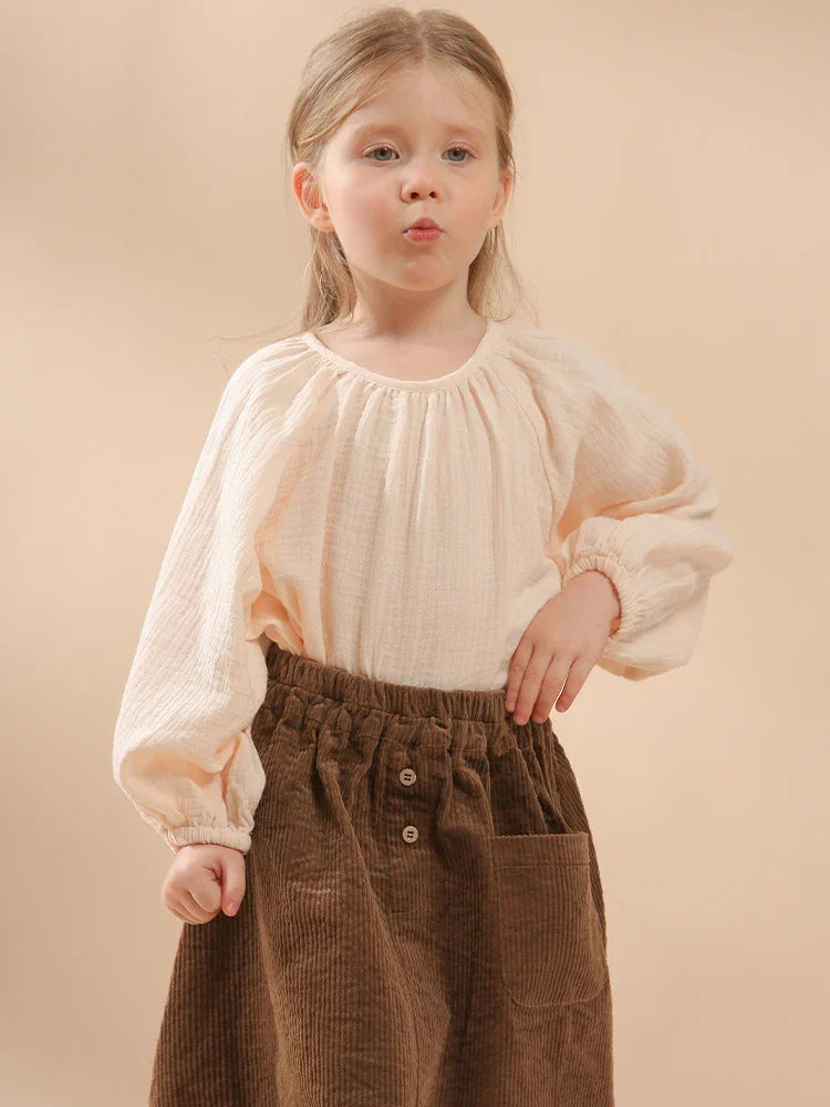 Young girl in cream blouse and brown corduroy skirt posing against beige background