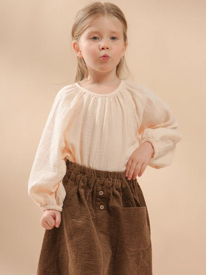 Young girl in cream blouse and brown corduroy skirt posing against beige background