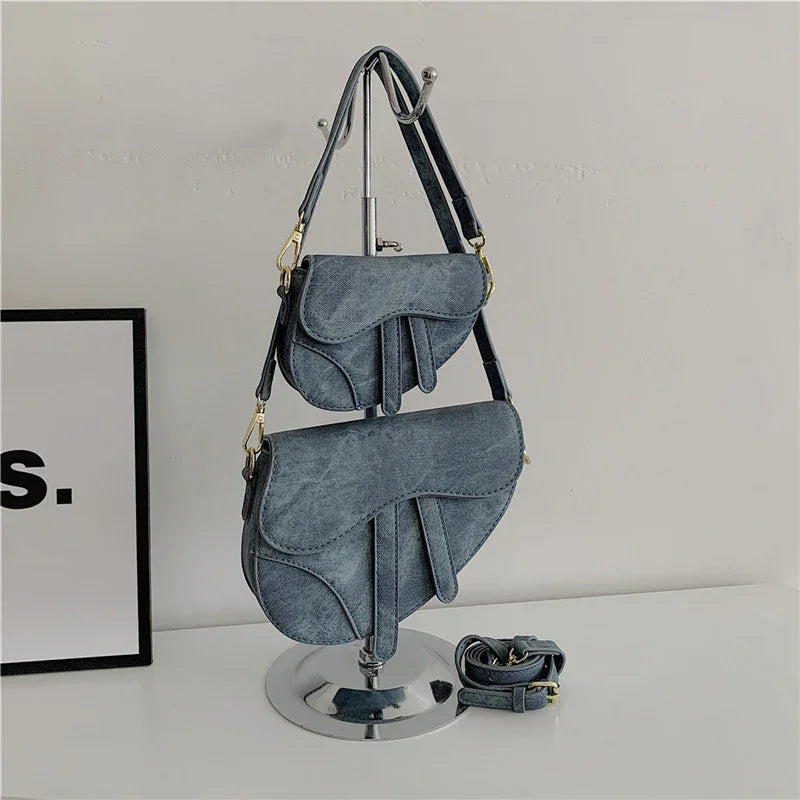 Two gray suede saddle bags displayed on a metal stand with adjustable straps on a white surface