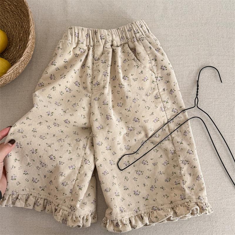 Beige cotton shorts with small purple floral pattern and ruffled hems on a light fabric surface with black wire hanger and woven basket