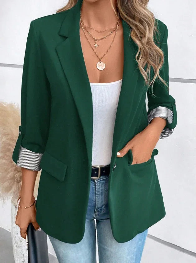 Woman wearing dark green blazer with rolled striped cuffs, white tank top, layered gold necklaces, and light blue jeans
