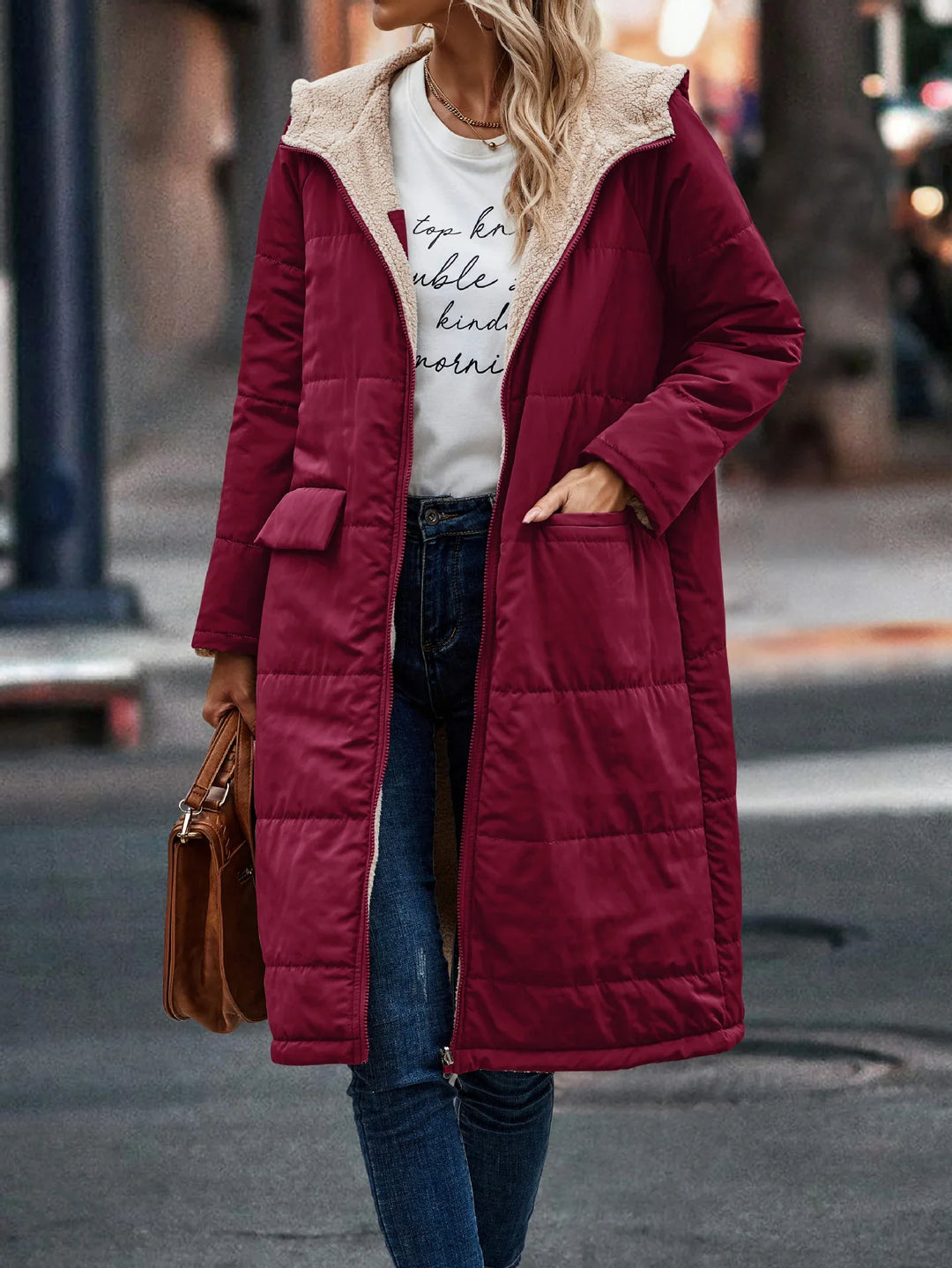 Woman wearing long burgundy quilted winter coat with sherpa lining, white printed shirt, skinny jeans, and brown handbag