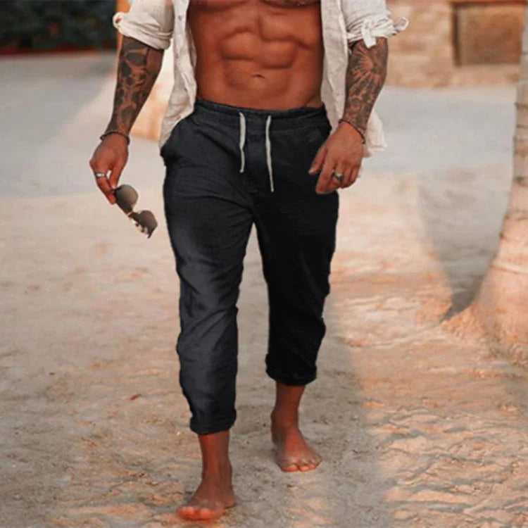 Muscular man with tattoos wearing black joggers and white shirt, walking barefoot on sandy beach