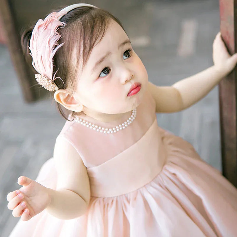 cute toddler girl in pink dress with pearl necklace and feather headband looking up
