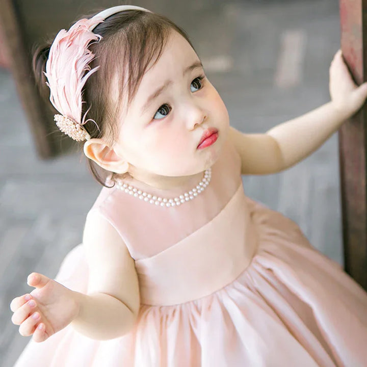 cute toddler girl in pink dress with pearl necklace and feather headband looking up
