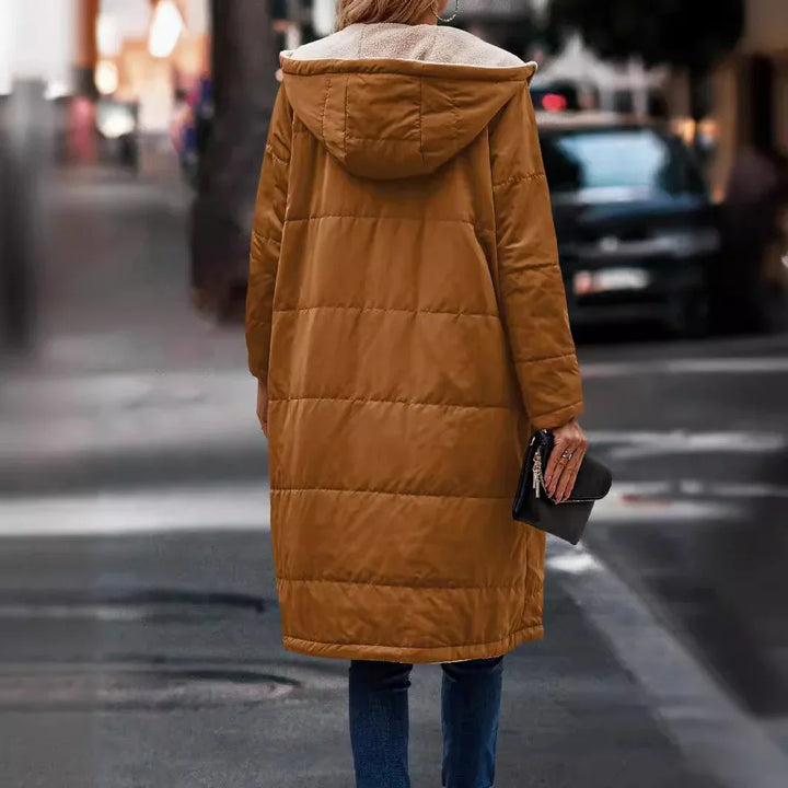 Woman wearing a long brown hooded puffer coat walking on city street holding black clutch