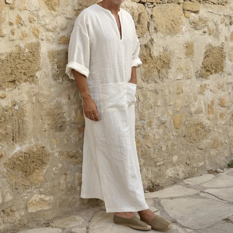 Man wearing white linen robe with rolled sleeves and beige espadrilles standing against stone wall