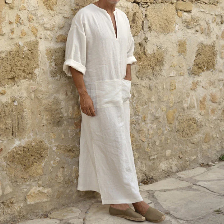 Man wearing white linen robe with rolled sleeves and beige espadrilles standing against stone wall