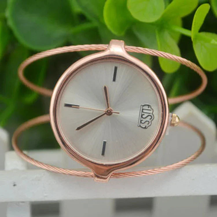 Rose gold wristwatch with minimalist dial and braided band on a white fence with green leaves background
