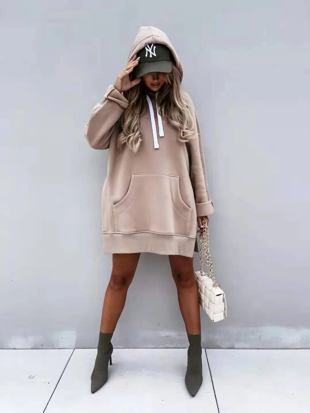 Woman in beige oversized hoodie dress, green cap, and green ankle boots holding white quilted handbag