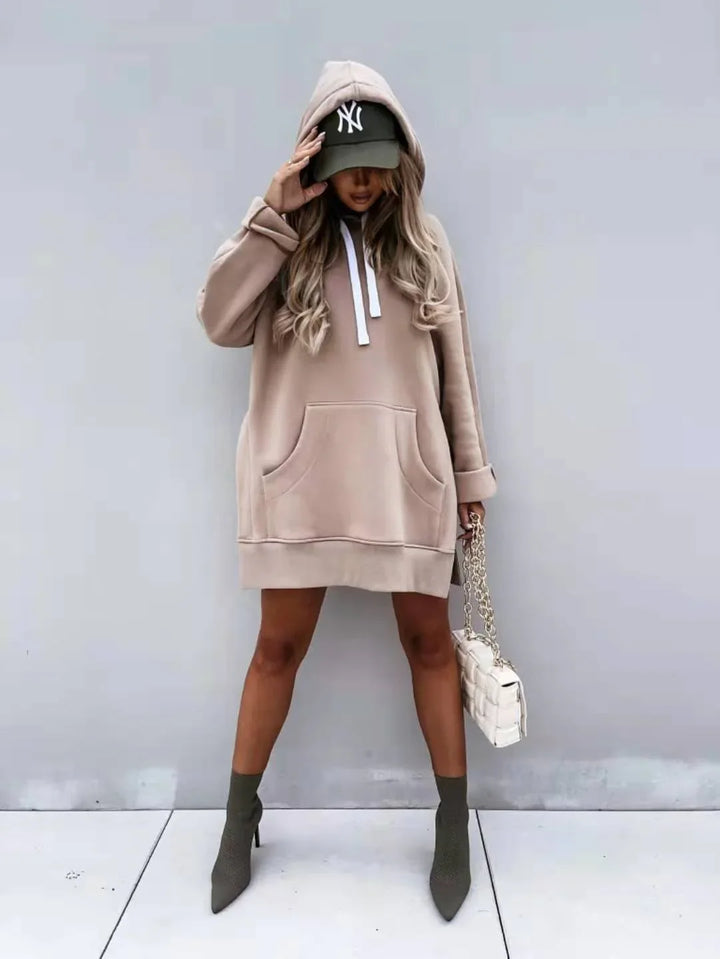 Woman in beige oversized hoodie dress, green cap, and green ankle boots holding white quilted handbag