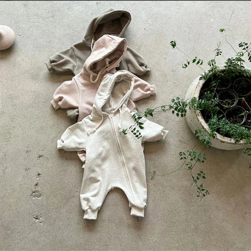 Three cozy baby hooded zip-up jumpsuits in cream, blush pink, and olive green on concrete floor next to potted plant