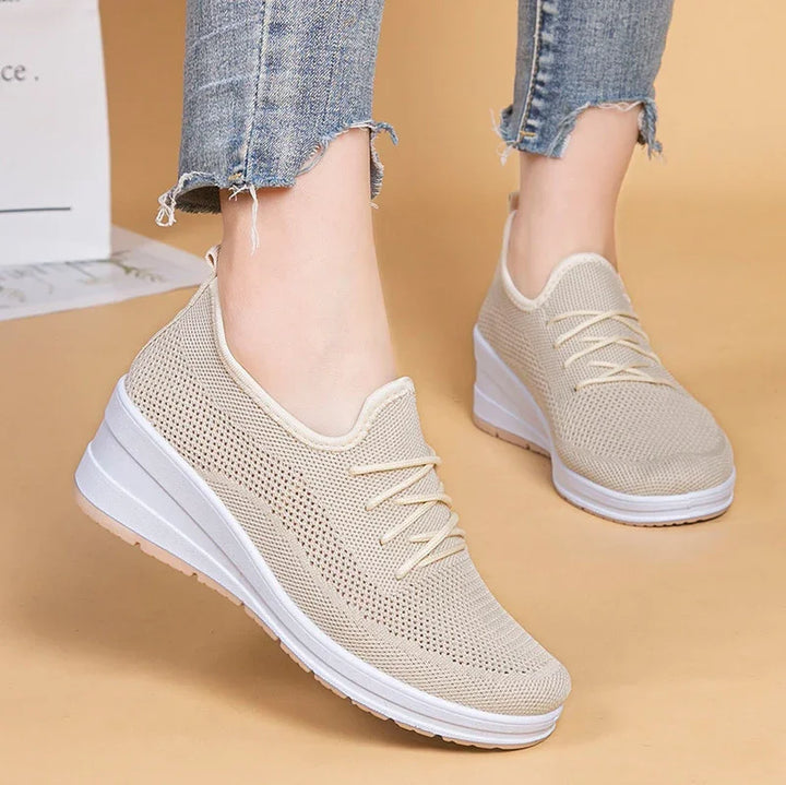Beige knitted sneakers on feet with white platform soles, worn with frayed blue jeans on tan background