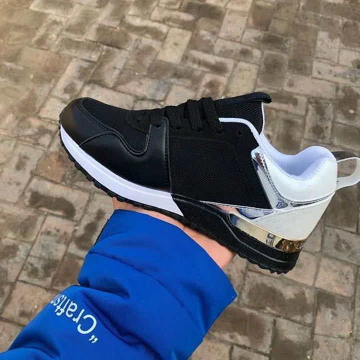 Black and white athletic sneaker with metallic silver heel detail held over outdoor brick pavement