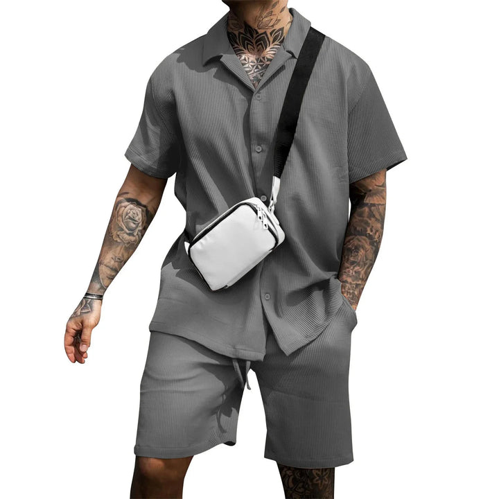 Man wearing gray ribbed short-sleeve shirt and shorts with white crossbody bag and visible tattoos
