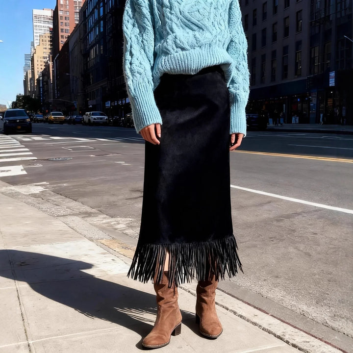 Woman wearing light blue cable knit sweater, black fringe midi skirt, and brown suede boots on city sidewalk