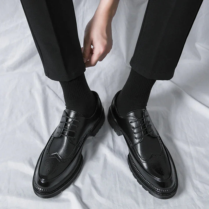 Person wearing black leather brogue dress shoes with black socks and black pants on white fabric