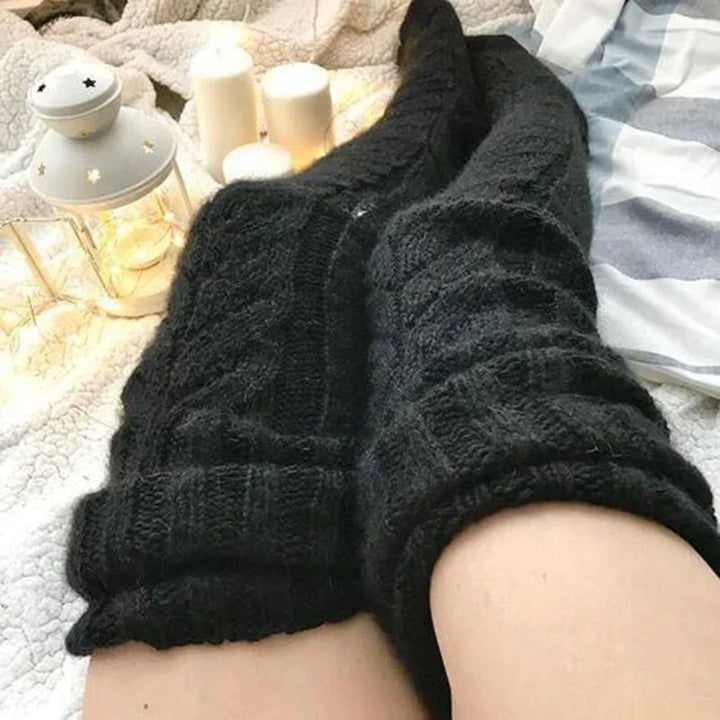 Cozy black knitted socks worn on legs with white candles and a white lantern in soft bedroom setting