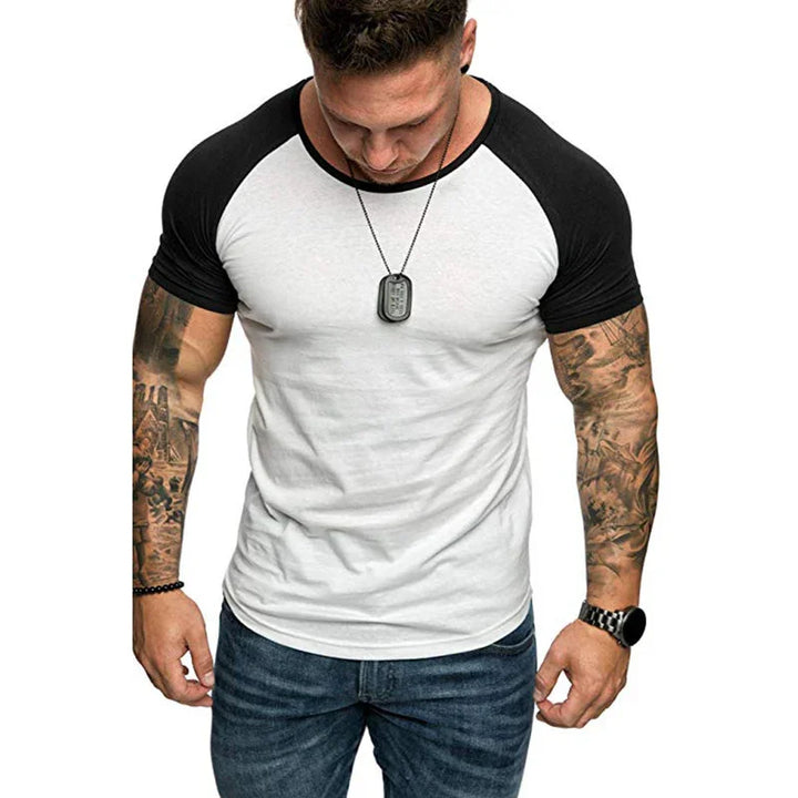 muscular man wearing white and black raglan sleeve t-shirt, blue jeans, and dog tag necklace with tattooed arms