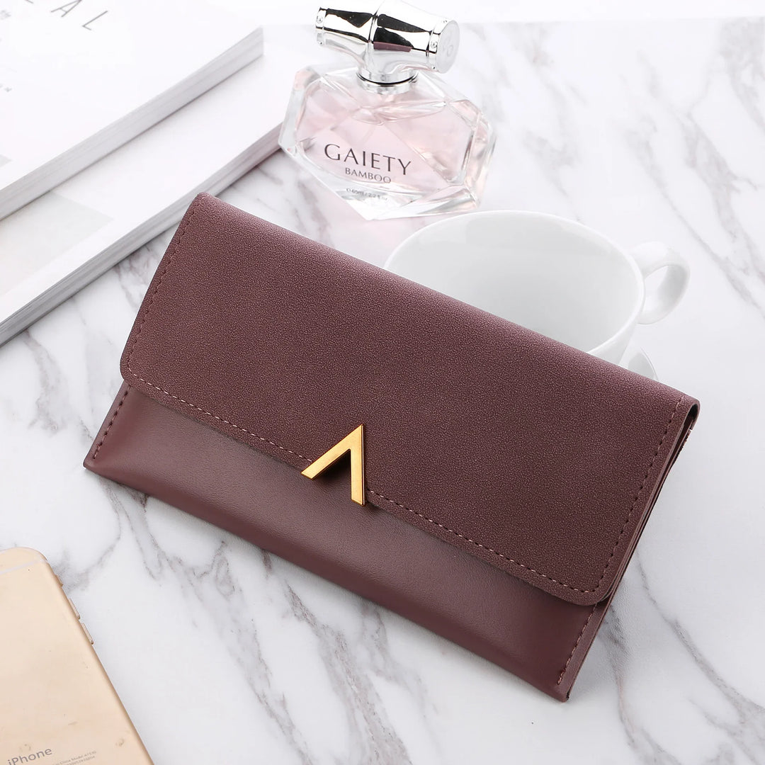 Elegant brown leather wallet with gold V clasp on marble surface next to perfume bottle and white cup