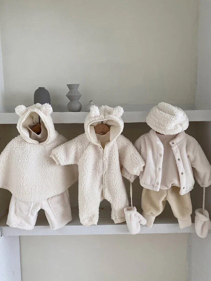 Three cream-colored baby winter outfits including bear-ear hooded jackets, mittens, and cozy pants displayed on a minimalist shelf