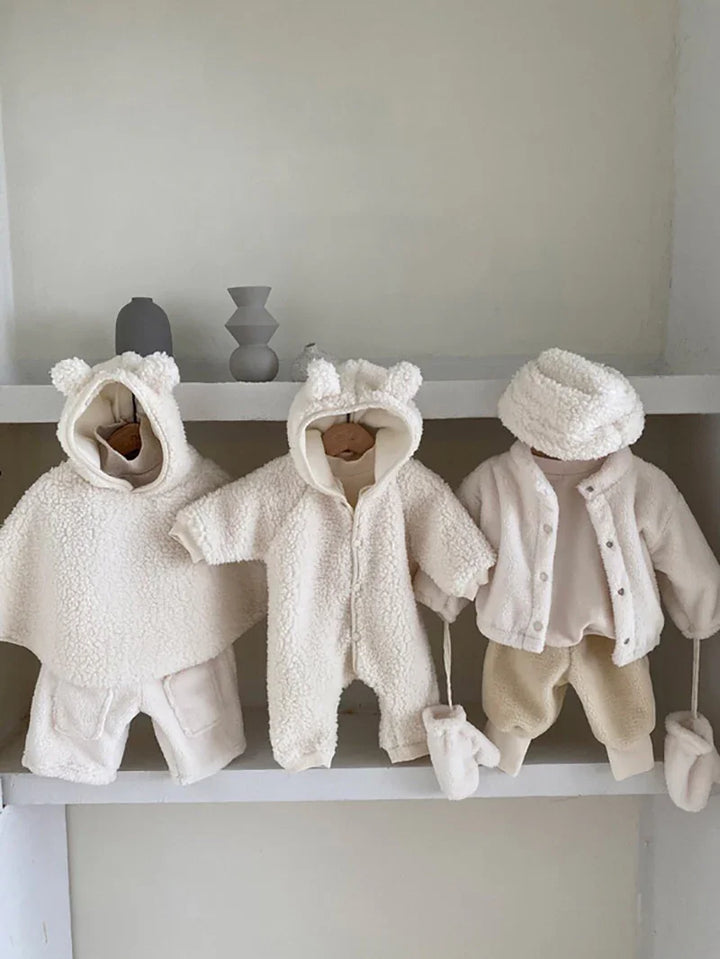 Three cream-colored baby winter outfits including bear-ear hooded jackets, mittens, and cozy pants displayed on a minimalist shelf