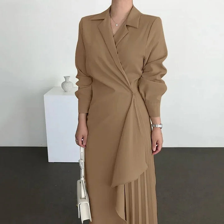 Woman wearing camel brown wrap dress with pleated skirt detail, holding white handbag against minimal background