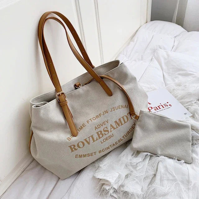 Beige canvas tote bag with brown leather straps and printed text, accompanied by a matching pouch on a white fabric surface