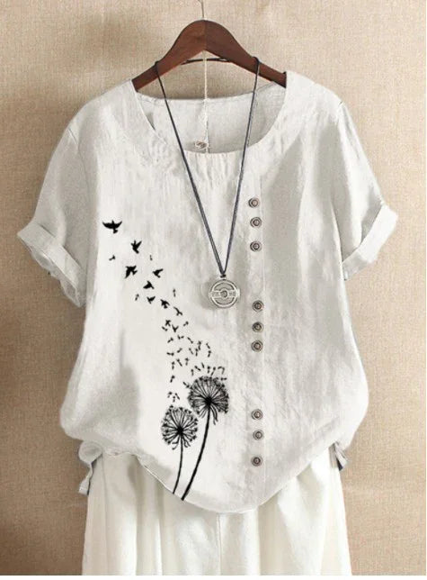 White short-sleeve blouse with black dandelion and flying birds print, button details, and a necklace on wooden hanger