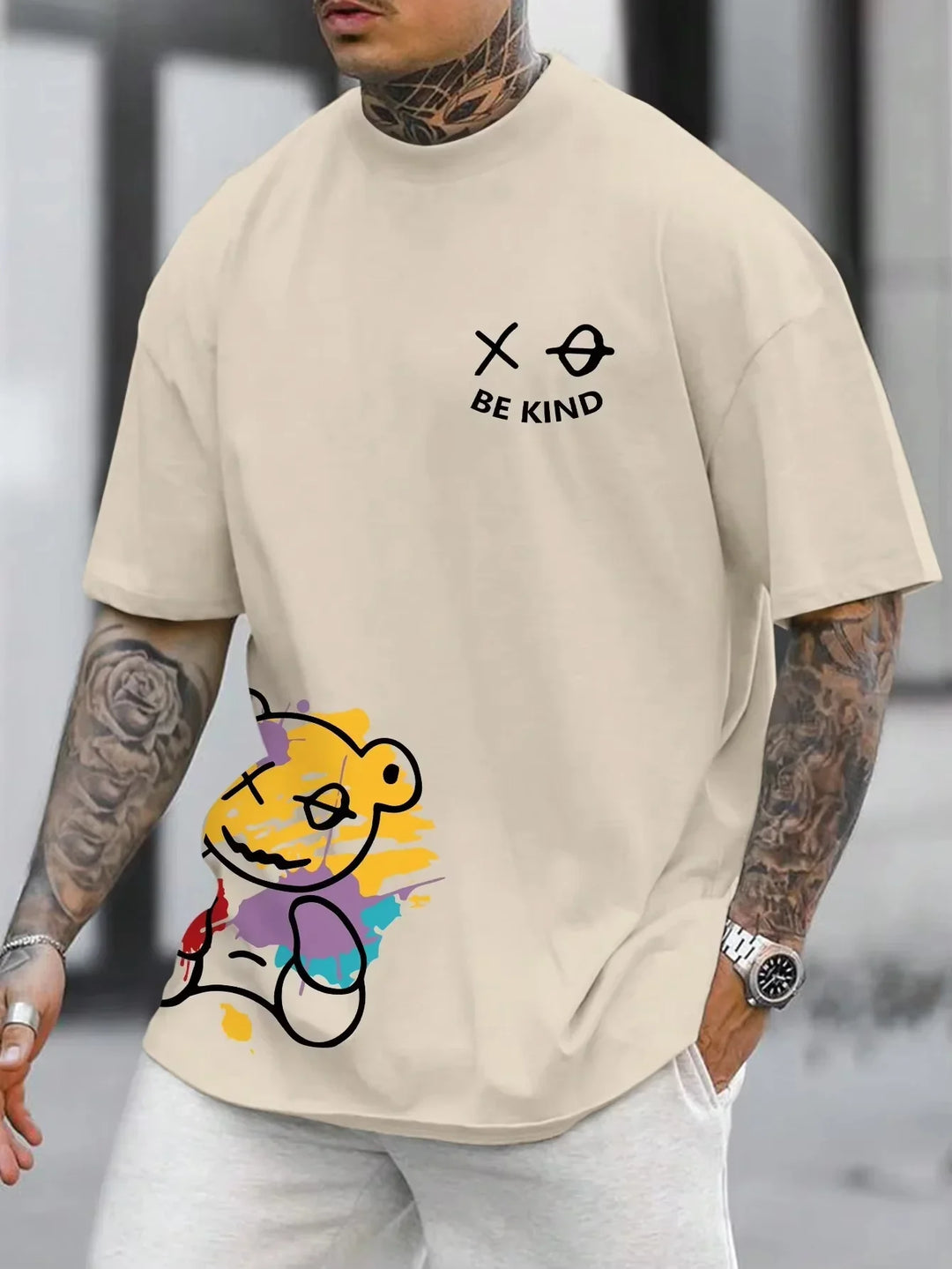 Beige oversized t-shirt with colorful abstract teddy bear and black 'BE KIND' text worn by tattooed man