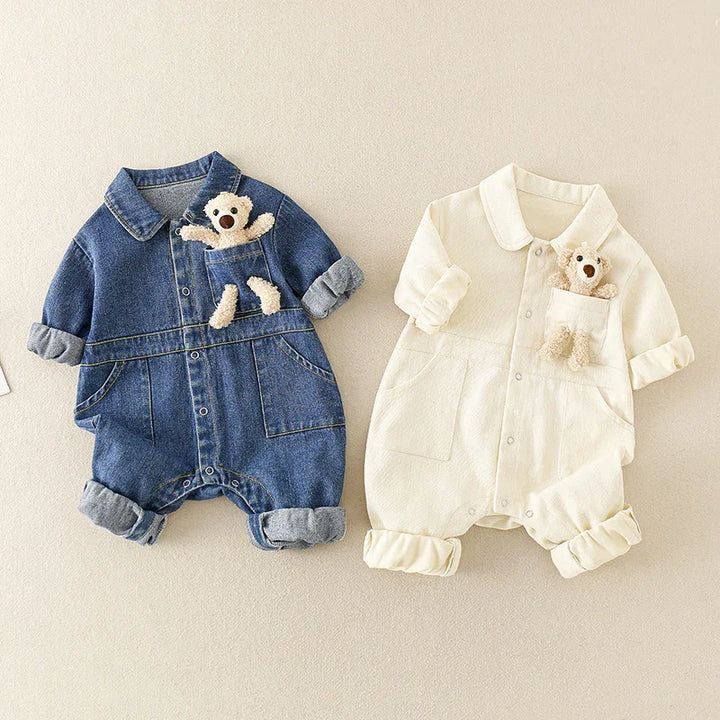 Two baby rompers with rolled sleeves and legs, one denim blue and one cream, each with a plush teddy bear in chest pocket