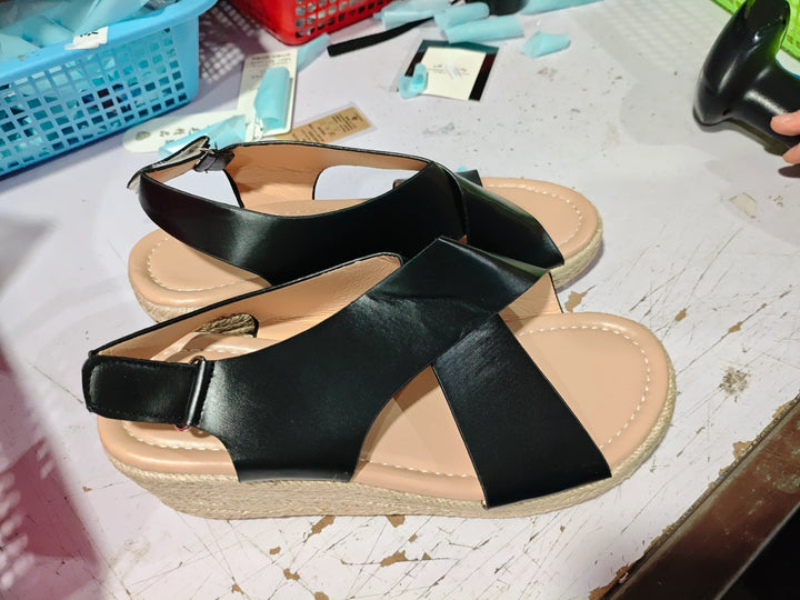 Pair of black leather sandals with beige cushioned soles on a white surface with baskets and tools