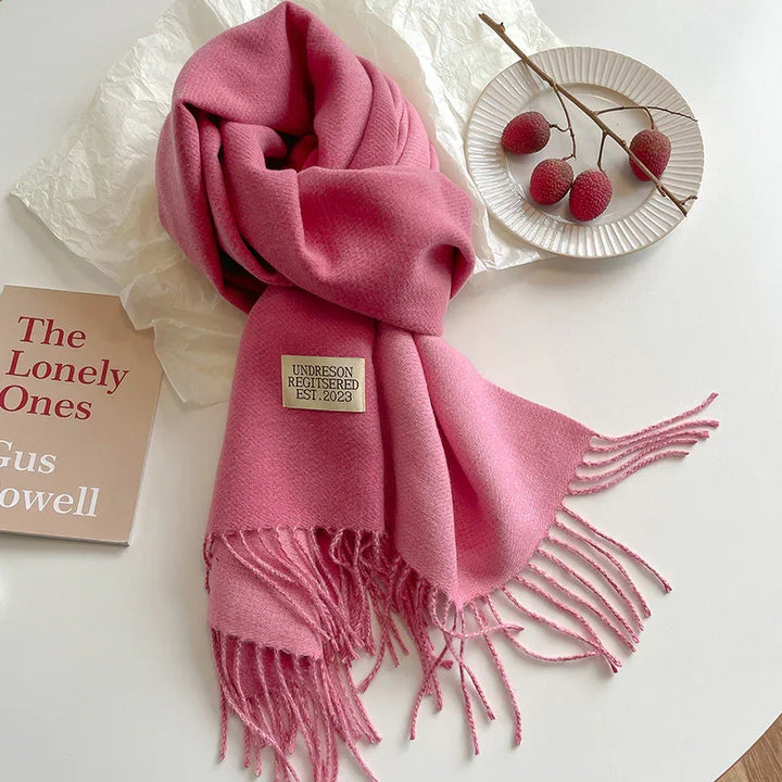 Soft pink fringed scarf with label on white table next to book and plate with red berries