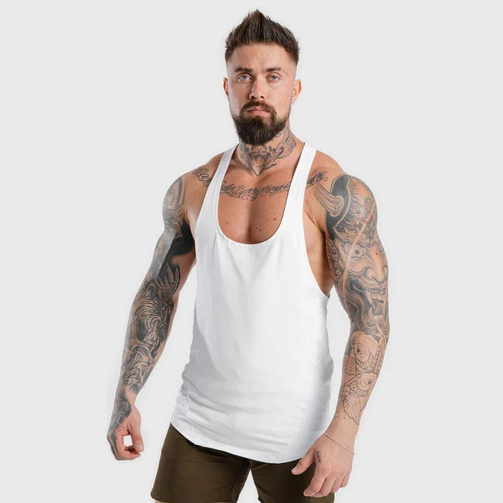 Muscular tattooed man wearing white gym tank top and brown shorts against plain background