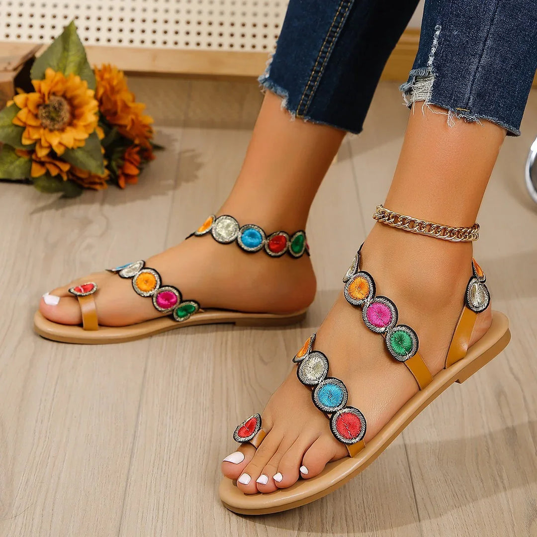 Feet wearing colorful embroidered flat sandals paired with gold anklet and denim jeans on wooden floor