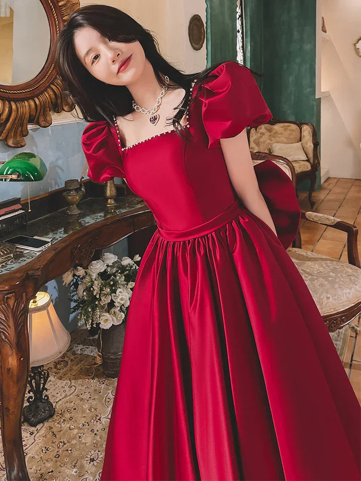 Woman in a red satin puff-sleeve ball gown standing indoors near an ornate wooden table with flowers