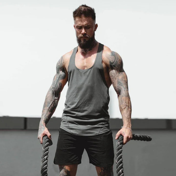 Muscular tattooed man in gray tank top and black shorts holding battle ropes in outdoor workout