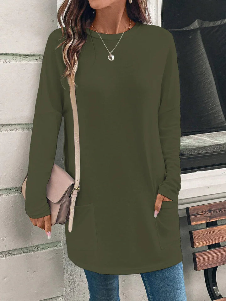 Woman wearing olive green long-sleeve tunic sweater with front pockets, carrying beige crossbody bag