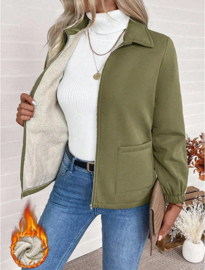 Woman wearing olive green fleece-lined jacket over white turtleneck with blue jeans holding clutch