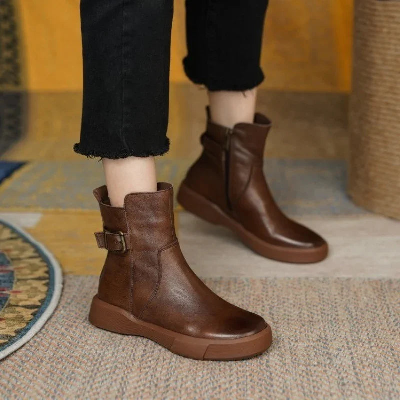 Brown leather ankle boots with side buckle and zipper worn with black frayed hem pants on a textured rug