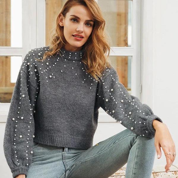 Woman wearing stylish gray pearl-embellished knit sweater and light blue jeans sitting indoors