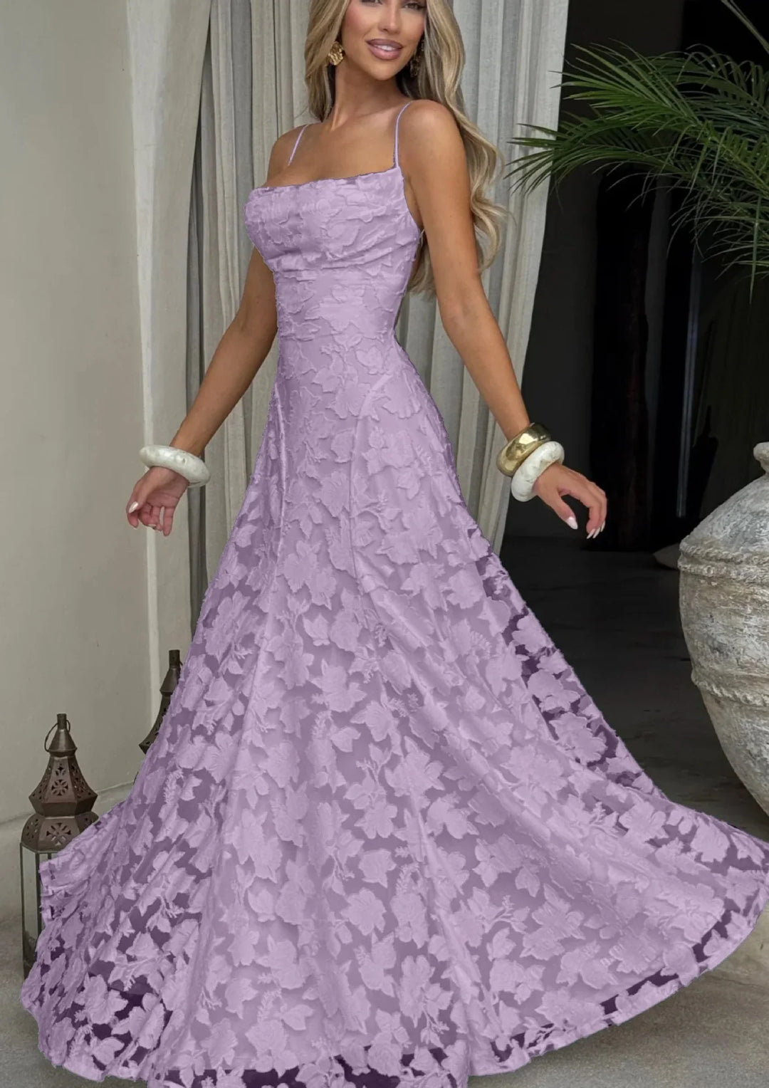 Woman in a lilac floral lace evening gown with spaghetti straps, posing indoors with bracelets