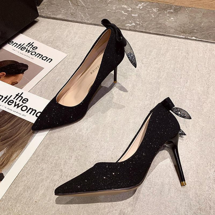 Pair of elegant black glitter high heel pumps with bows on back, displayed on stone floor near magazines