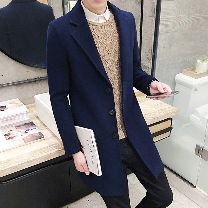 Man wearing navy blue overcoat and beige cable knit sweater holding book and phone indoors