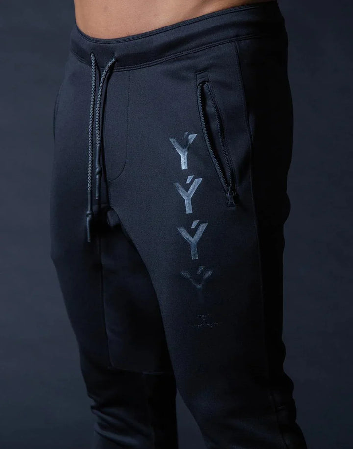 Close-up of black jogger pants with zipper pocket and Y-shaped logo print on leg against dark background