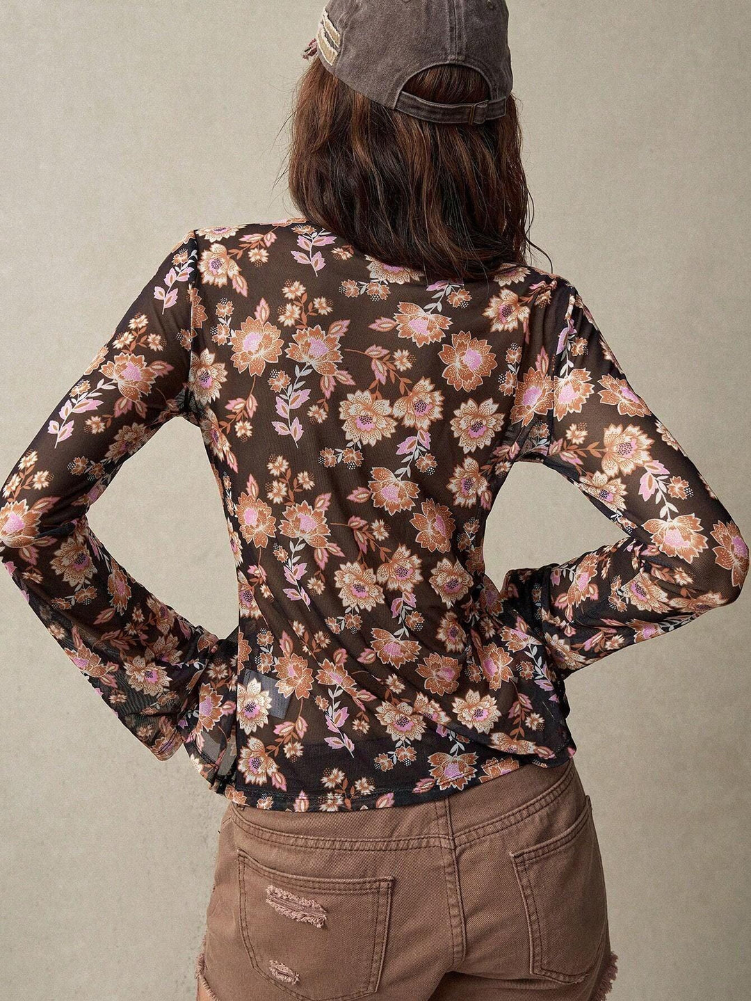 Back view of woman wearing sheer black floral long-sleeve top and distressed brown shorts with a gray cap