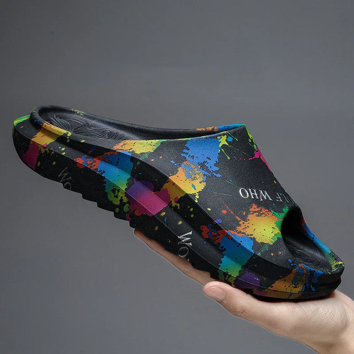 Hand holding black slide sandal with colorful paint splatter design and textured sole